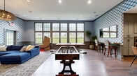 This thoughtfully designed single-floor amenity space offers a compelling blend of entertainment, socializing, and functionality. The interiors are immediately distinguished by striking blue-and-white scallop-patterned wallcoverings that extend throughout, complemented by wainscoting panels in a warm gray tone and rich hardwood flooring underfoot.

The expansive game and lounge area serves as the centerpiece of the space. A foosball table and ping-pong table anchor the room, while a plush navy blue sectional sofa and a round leather ottoman create a comfortable viewing and lounging zone. A large flat-screen display is mounted on the feature wall, and generous windows along the perimeter flood the space with natural light while offering views of a manicured green lawn.

Adjacent to the lounge, a well-appointed kitchen is outfitted with gray shaker-style cabinetry, dark granite countertops, a bold navy hexagonal tile backsplash, stainless steel appliances including a double wall oven, and a deep undermount sink. A central island with bar seating provides additional prep and gathering space, while pendant lighting adds warmth overhead.

A dedicated dining area and hallway complete the layout, ensuring smooth flow throughout. Tall indoor plants add natural accents, enhancing the polished, welcoming ambiance of the entire space.