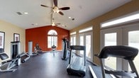 This single-floor fitness space is designed to support a comprehensive workout experience in a bright, open environment. The room features vaulted ceilings that lend a sense of openness, complemented by two ceiling fans that ensure consistent air circulation throughout. Recessed lighting adds to the well-lit atmosphere alongside generous natural light streaming in through multiple large windows and glass-paneled doors along one wall.

The interior design makes a bold visual statement with a deep terra cotta-orange accent wall on one end, contrasting with warm yellow-toned walls on the sides. An arched window above the accent wall adds an architectural focal point and draws in additional light. The flooring is a durable dark rubberized surface, practical for a fitness setting and easy to maintain.

The space is equipped with cardio machines, including treadmills and an elliptical-style stepper, as well as strength training equipment such as a cable machine and a weight chair. The layout provides adequate spacing between equipment for safe and comfortable use. The glass doors offer views to the outdoors, creating an inviting and energizing atmosphere for workouts.