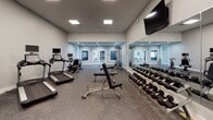 Located at 3600 Harry S Truman Blvd in Saint Charles, Missouri, this fitness center offers a comprehensive workout environment spread across a single floor with distinct zones designed to support a variety of training styles. The cardio area is outfitted with treadmills, elliptical machines, and a stationary bike, each equipped with individual monitor screens, all set on durable rubber flooring beneath a drop-panel ceiling with recessed lighting. A dedicated strength training section features a multi-station cable machine, adjustable benches, and a full rack of dumbbells neatly arranged along a mirrored wall, with a boldly designed geometric accent wall adding visual energy to the space. The functional training studio is a standout area, featuring herringbone-patterned carpet tile flooring, ceiling fans, large windows that bring in natural light, and wall-mounted suspension training rigs alongside resistance bands and stability balls. Full-length mirrors and a wall-mounted mat complete the setup for stretching and bodyweight work. French doors connect the zones seamlessly, and the clean, neutral color palette throughout creates a bright, motivating atmosphere. The fitness center is thoughtfully designed to accommodate cardio, strength, and functional training all in one cohesive facility.