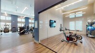 Located at 12609 Matisse Cir in New Port Richey, FL, this modern fitness center is designed to deliver a comprehensive workout experience within a bright, open, and thoughtfully appointed environment. The facility spans a single floor and is distinguished by its soaring double-height ceilings, which lend an expansive, airy atmosphere throughout the main exercise area.

The primary workout floor is lined with floor-to-ceiling windows that flood the space with natural light and offer views of the surrounding palm-dotted Florida landscape. The room is outfitted with a full suite of Life Fitness equipment, including treadmills and ellipticals with integrated display screens, a cable functional trainer, a multi-station strength machine, a leg press, free-weight benches, and a well-stocked dumbbell rack. Full-length mirrors line one wall, enhancing the sense of space and supporting proper form during training.

A dedicated spin studio is separated from the main floor by sleek glass sliding doors, providing a semi-private space for cycling workouts. The studio features spin bikes equipped with performance monitors, a wall-mounted television, and wood-look flooring that sets it apart from the rubberized flooring of the main area.

Throughout the facility, the design is cohesive and contemporary, featuring a muted blue-gray color palette, linear pendant lighting, recessed ceiling fixtures, and vertical slatted wall panels that add visual texture. The overall ambiance is clean, professional, and inviting.