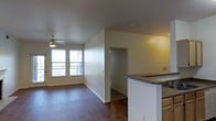 This single-floor space showcases an open-concept design that seamlessly connects the kitchen and living areas. The kitchen features a wraparound peninsula with a dark countertop, providing both workspace and a natural divider between the cooking and living zones. A built-in sink is visible along the peninsula, and cabinetry lines the kitchen walls, offering ample storage.

The living area is finished with rich, dark hardwood-style flooring that extends throughout the space, adding warmth and visual continuity. Two side-by-side windows allow natural light to fill the room, complementing the ceiling fan with a light fixture mounted above. The neutral, cream-toned walls keep the space feeling open and versatile.

A doorway visible in the background leads to an additional room or hallway, suggesting further functional areas beyond the main living space. The layout is efficient and well-organized, with clearly defined zones for cooking and relaxing. Overall, the space presents a clean, practical interior suited for everyday living.