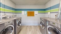 Located at 1808 Kuser Rd in Trenton, NJ, this communal laundry room is a compact, efficiently organized space designed to serve its users with practicality and a welcoming aesthetic. The room's most distinctive design feature is its painted cinder block walls, adorned with bold horizontal stripes in teal, lime green, and gray — adding a vibrant, modern touch to an otherwise utilitarian space.

The room is equipped with multiple LG commercial-grade front-loading washing and drying machines, positioned along opposing walls to maximize the use of floor space. A white folding table with hairpin legs provides a convenient surface for sorting and folding laundry, complementing the functional layout. Exposed plumbing and ventilation ductwork run along the walls and ceiling, reflecting the room's straightforward, no-frills infrastructure.

Light wood-look flooring runs throughout, lending a clean and contemporary feel underfoot. Natural light filters in through a window and a windowed exterior door, which also provides direct outdoor access. A bulletin board mounted on the rear wall serves as an informational hub for residents. The overall ambiance is tidy, bright, and purpose-driven, making this laundry room a practical and pleasant shared amenity.
