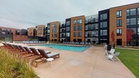 This expansive outdoor patio area is designed to serve as a premier amenity space, combining relaxation and social gathering in a well-appointed setting. The centerpiece is a large rectangular swimming pool with clear blue water, surrounded by a broad concrete deck furnished with numerous cushioned lounge chairs and side tables, providing ample space for sunbathing and leisure. The pool area is framed by the backdrop of a modern multi-story building featuring a contemporary exterior of warm brick tones and dark navy panels with large windows.

Adjacent to the pool deck is a covered outdoor pavilion, anchored by a sleek tensile shade sail and a flat roof structure supported by steel columns. This sheltered area houses a communal dining zone with long tables and chairs, alongside an outdoor kitchen or bar counter, making it well-suited for entertaining and casual dining. The pavilion connects directly to a glass-walled interior amenity space, allowing for a seamless indoor-outdoor experience.

Ornamental grasses and native plantings are integrated throughout the patio to soften the hardscape and add natural texture. The overall design reflects a modern aesthetic, with clean lines, durable materials, and thoughtful zoning that accommodates both active and relaxed outdoor living.