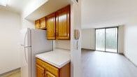 This well-organized single-floor residence offers a practical and comfortable layout comprising a living room, bedroom, kitchen, bathroom, and entrance. The living room is bright and inviting, featuring light hardwood-style flooring and a large sliding glass door dressed with vertical blinds that allows abundant natural light and provides access to an outdoor view. The galley-style kitchen is outfitted with warm honey-toned oak cabinetry — both upper and lower — paired with white countertops, a full-size refrigerator, stove, and sink, creating a fully functional cooking space with ample storage. An intercom unit is conveniently positioned at the kitchen's entrance. The bedroom offers a cozy atmosphere with plush carpeting, neutral walls, and a mirrored sliding-door closet that adds depth and storage. The bathroom is clean and well-appointed, featuring a white vanity with under-cabinet storage, a wall mirror, and a combined shower and bathtub with hardwood-style flooring tying the space together. Throughout the residence, the consistent use of light tones, neutral walls, and natural light creates a clean, airy ambiance that maximizes the sense of space within this thoughtfully designed home.