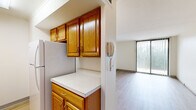 This well-organized single-floor residence offers a practical and comfortable layout comprising a living room, bedroom, kitchen, bathroom, and entrance. The living room is bright and inviting, featuring light hardwood-style flooring and a large sliding glass door dressed with vertical blinds that allows abundant natural light and provides access to an outdoor view. The galley-style kitchen is outfitted with warm honey-toned oak cabinetry — both upper and lower — paired with white countertops, a full-size refrigerator, stove, and sink, creating a fully functional cooking space with ample storage. An intercom unit is conveniently positioned at the kitchen's entrance. The bedroom offers a cozy atmosphere with plush carpeting, neutral walls, and a mirrored sliding-door closet that adds depth and storage. The bathroom is clean and well-appointed, featuring a white vanity with under-cabinet storage, a wall mirror, and a combined shower and bathtub with hardwood-style flooring tying the space together. Throughout the residence, the consistent use of light tones, neutral walls, and natural light creates a clean, airy ambiance that maximizes the sense of space within this thoughtfully designed home.