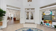 Located at 4688 New Broad St in Orlando, FL, this single-story community clubhouse makes a striking first impression with its dramatic rotunda-style entry foyer. Soaring double-height ceilings clad in crisp white shiplap paneling frame a set of elegant glass-paneled double doors, while a decorative medallion above the entrance and a bold area rug anchoring the tiled floor add refined character to the arrival experience.

The interior opens into a series of well-appointed gathering spaces filled with natural light. Floor-to-ceiling windows and French doors line the lounge and seating areas, offering lush outdoor views of the surrounding landscaping. The lounge features coffered ceilings with lantern-style pendant lighting, neutral toned walls, and comfortable seating arrangements that create an inviting atmosphere for socializing.

A dedicated dining and kitchen area provides a full suite of stainless steel appliances, dark wood cabinetry, white subway tile backsplash, and a large communal dining table beneath a trio of dramatic metallic cone pendant lights. A bistro-style seating nook with high-top tables and bar stools sits beside oversized windows, bathing the space in warm natural sunlight. Throughout the clubhouse, light wood-look tile flooring, carefully curated furnishings, and lush potted greenery contribute to a bright, polished, and welcoming ambiance.