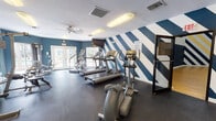 Located at 2654 N McMullen Booth Rd in Clearwater, Florida, this single-floor fitness and recreation facility offers a well-rounded environment for both cardio training and competitive sport. The fitness area is designed with an energetic aesthetic, highlighted by bold navy and white diagonal striped accent walls. This space is outfitted with a range of cardio equipment including treadmills, elliptical trainers, and a stair climber, as well as strength training machines and free weight options. Rubber flooring provides a safe, durable surface throughout, and a ceiling fan along with ample overhead lighting ensure a comfortable workout environment. French doors offer a connection to the outdoors and bring in natural light. A secondary fitness area provides additional strength and cross-training machines, with direct access to the adjacent basketball court. The indoor basketball court is a standout feature of the facility, boasting soaring ceilings, regulation hardwood flooring with clearly marked court lines, and glass backboard hoops at each end. Rows of high clerestory windows allow natural light to filter in, brightening the space. The court walls carry the same bold blue, white, and gold diagonal stripe motif, creating a cohesive and visually dynamic atmosphere throughout the entire facility.