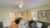 This single-floor residence features an open-concept layout that seamlessly connects the living and dining areas, creating a cohesive and functional living environment. The living area is anchored by a ceiling fan with a light fixture, which provides both practical ventilation and ambient lighting. A textured popcorn ceiling adds a classic touch overhead, while warm beige walls and light hardwood-style flooring contribute to an inviting atmosphere throughout.

The living space is furnished with a patterned accent chair and a plush sofa, complemented by a dark-toned console with a table lamp, offering a cozy and well-lit corner. Adjacent to the living area, the dining space accommodates a dark wood table and matching chairs, centered beneath a recessed skylight that introduces a note of natural light. Framed artwork adorns the walls, adding a personal character to the space.

A hallway visible in the background suggests additional rooms beyond the main living area. Overall, the space presents a straightforward, comfortable layout with consistent neutral tones and practical design choices throughout.