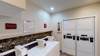 Located at 2904 Clairemont in San Diego, this shared laundry facility is a clean, functional, and well-maintained space designed to serve the needs of its users efficiently. The room features a row of commercial-grade top-loading washing machines equipped with digital controls, positioned along a wall adorned with a decorative mosaic tile backsplash that adds a touch of style to the utilitarian space. Opposite the washers, two sets of stacked dryers provide ample drying capacity, each unit featuring easy-to-use digital interfaces. Recessed lighting in the smooth ceiling keeps the room bright and welcoming. The floor is finished with durable slate-style tile in warm earth tones, contributing to a clean and practical aesthetic. A spacious laminate folding counter runs along one wall, offering convenient space for sorting and folding laundry. A wall-mounted clothing rack provides additional functionality for hanging garments. Safety is addressed with a clearly visible fire extinguisher cabinet. Posted signage from WashTek provides usage guidelines, and a security camera ensures the safety of users. The space is accessed through a glass-paneled entry door and includes a utility sink, making this laundry room a comprehensive and user-friendly facility.