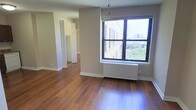 This single-floor residence offers a clean, functional layout highlighted by warm, rich hardwood flooring that extends throughout the main living areas and into the adjoining spaces. The living area is bathed in natural light from a large window framing an elevated view of the surrounding cityscape, including a lush tree canopy and distant urban skyline — a standout feature that adds depth and openness to the interior. A radiator beneath the window provides efficient heating, complementing the space's practical design.

The neutral, warm-toned walls create a versatile backdrop well-suited to a range of furnishing styles. The open corridor leads to additional rooms, each maintaining the same hardwood flooring and consistent aesthetic. Closet space is notably well-appointed, with bifold-door closets offering generous storage solutions in the adjoining areas.

White trim and baseboards throughout provide a crisp contrast against the warm tones of the floors and walls, lending the space a polished, tidy character. The overall ambiance is bright, airy, and understated — ideal for those seeking a comfortable, well-lit living environment with appealing urban views and thoughtful storage throughout.