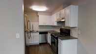 This single-floor space is thoughtfully designed with a clean, modern aesthetic that balances functionality and style. The kitchen serves as a standout feature, showcasing crisp white shaker-style cabinetry that extends to the upper walls, providing generous storage throughout. Dark charcoal countertops offer a striking contrast against the white cabinetry, while a classic white subway tile backsplash ties the design together with a timeless touch. The kitchen is equipped with stainless steel appliances, including a refrigerator and dishwasher, complemented by a black electric range, and an integrated range hood above. A stainless steel sink with a modern pull-down faucet is neatly positioned beneath the cabinetry, adding to the space's practical layout. The kitchen floor features rich, dark hardwood-style flooring that adds warmth and depth to the space. Overhead, a large flush-mount circular light fixture provides bright, even illumination throughout the kitchen. The L-shaped layout of the kitchen maximizes efficiency, making it both a practical and visually appealing area. The overall ambiance is fresh, clean, and move-in ready, reflecting a contemporary design sensibility throughout.