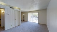This single-floor residence offers a straightforward and functional layout designed for comfortable everyday living. The main living area is bright and open, anchored by large sliding glass doors fitted with vertical blinds that allow generous natural light to fill the room while providing a view of the lush greenery outside. The space is finished with plush, neutral-toned carpet flooring that adds warmth underfoot, while the textured popcorn ceiling lends a classic residential character. The walls are painted in a clean, light neutral tone that keeps the interior feeling airy and welcoming. Adjacent to the living area, the kitchen is accessible through a defined opening, revealing white cabinetry and matching white appliances, including a range, creating a cohesive and tidy cooking space. A doorway leading to additional rooms is visible from the main living space, suggesting a connected and navigable floor plan. The overall ambiance of the residence is calm and uncluttered, making it a versatile space that can be tailored to a variety of furnishing styles and personal preferences. The abundance of natural light and the outdoor view contribute to an inviting atmosphere throughout the home.