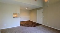 This single-floor space offers a clean, open layout designed for functional living. The main living area combines two distinct flooring materials — rich, dark hardwood-style planks near the entryway transitioning into soft, plush carpeting in the main lounge area — creating defined zones within the open floor plan. Neutral beige walls and crisp white baseboards contribute to a bright, cohesive aesthetic throughout.

A white panel entry door with a deadbolt provides a welcoming focal point at the front of the space, while recessed lighting in the ceiling ensures consistent illumination. The textured ceiling adds subtle architectural character to the otherwise streamlined interior.

Adjacent to the living area, a kitchen pass-through with a granite-style countertop provides a convenient connection between the cooking and living spaces, allowing for easy interaction between rooms. The raised counter also serves as a visual divider without fully closing off the kitchen.

Overall, the space maintains a straightforward, well-maintained atmosphere with a neutral palette and practical design choices that provide a versatile and comfortable environment.