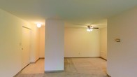 This single-floor space presents an open, functional layout defined by clean lines and a neutral color palette. The main living area is finished with wall-to-wall carpet in a light, neutral tone, contributing to a quiet and comfortable atmosphere. A central structural column divides the space, creating a natural visual separation between areas while maintaining an open feel throughout. The ceiling features a ceiling fan with an integrated multi-bulb light fixture, providing both air circulation and ambient lighting. An additional flush-mounted light fixture is visible in the adjoining area, ensuring consistent illumination throughout. The walls are painted in a soft, warm off-white, complemented by standard baseboards that run along the perimeter. The layout suggests a generous living area with room for flexible furniture arrangements. Overall, the space offers a clean, move-in-ready foundation with practical features suited for everyday living.