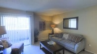 This single-floor residence centers around a comfortable living room that serves as the heart of the space. The living room features plush carpeting underfoot and neutral-toned walls that create a warm, welcoming atmosphere. A textured ceiling adds subtle visual interest overhead. Large vertical blinds frame a full-height sliding glass door, allowing generous natural light to fill the room while providing direct access to an outdoor area. The space is thoughtfully arranged with a cushioned sofa accented by striped and solid throw pillows, paired with a dark wood coffee table as a functional centerpiece. A decorative wall mirror with a dark frame reflects additional light, enhancing the sense of openness. A side table with a warm-toned lamp provides ambient lighting, complementing the natural light from the sliding door. Decorative wall art and a potted indoor plant add touches of color and life to the room. The overall ambiance is relaxed and comfortable, with a layout that balances practicality and style in a modest, well-appointed single-level living environment.