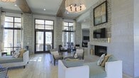 This single-floor space exemplifies refined design through a seamless blend of natural materials and contemporary elegance. The expansive main living area commands attention with its soaring ceilings and striking floor-to-ceiling stone accent walls that anchor the room with texture and warmth. A grand stone fireplace serves as a central focal point, topped with a mounted flat-screen television and framed by tasteful artwork above.

The space is generously furnished with a curated mix of plush seating, including upholstered sofas, patterned accent chairs, and a sleek central coffee table, all arranged to encourage conversation and comfort. Soft, neutral tones throughout the furnishings and finishes create a cohesive, calming palette.

Natural light pours in abundantly through floor-to-ceiling black-framed glass doors and windows, offering views of a bright outdoor area and blurring the boundary between interior and exterior. Light hardwood-style flooring adds warmth underfoot and complements the stone and neutral color scheme. An elegant chandelier overhead provides a polished finishing touch. The overall ambiance is one of casual sophistication — a welcoming, well-appointed space designed for both relaxation and social gathering.