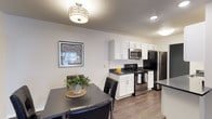 This single-floor space offers a clean, modern aesthetic with thoughtful design details throughout. The kitchen is a standout feature, showcasing crisp white shaker-style cabinetry paired with dark countertops that provide a striking contrast. A full suite of stainless steel appliances — including a range, over-the-range microwave, and refrigerator — equips the kitchen for everyday cooking needs. A classic white subway tile backsplash adds a timeless finishing touch to the cooking area. The flooring throughout is a warm-toned wood-look plank, lending continuity and a welcoming feel to the space. Adjacent to the kitchen, the dining area comfortably accommodates a dark-toned dining table and chairs, creating a natural space for meals and gathering. A decorative framed artwork adorns the wall, adding a touch of personality to the area. Overhead, a stylish semi-flush mount light fixture with a geometric design serves as a focal point and provides warm ambient lighting. Recessed lighting in the kitchen ensures the workspace remains well-lit. The textured ceiling and neutral wall tones contribute to a bright, understated atmosphere that is both practical and inviting.