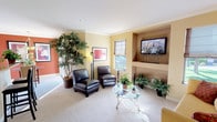 This single-floor space offers a warm and inviting atmosphere defined by its thoughtful layout and carefully curated design details. The living area serves as the focal point, featuring neutral carpeted flooring, soft yellow walls accented by a rich terracotta-toned feature wall, and elegant crown molding that adds a refined architectural touch to the interior.

A built-in recessed shelving unit provides both functional storage and a dedicated display space, currently housing a wall-mounted flat-screen television alongside an array of decorative potted plants and greenery. Natural light filters in through multiple windows dressed with fabric Roman shades, creating a bright yet comfortable ambiance throughout the day.

Seating is provided by a pair of sleek black leather accent chairs arranged around a round glass-top coffee table with an ornate metal base, encouraging casual conversation and relaxation. A tall, lush indoor tree positioned in the corner adds a striking natural element, enhancing the room's organic warmth. Complementing this is an assortment of smaller potted ferns and houseplants scattered throughout the space, bringing life and texture to the interior.

Overhead lighting is supplemented by a tall floor lamp, ensuring the space is well-lit at all times. Overall, the space presents a clean, comfortable, and well-appointed environment suitable for a variety of uses.