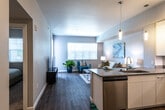 This thoughtfully designed single-floor residence offers a seamless blend of modern aesthetics and functional living. The open-concept layout connects the kitchen, dining, and living areas into one cohesive, light-filled space. Rich wood-look flooring flows throughout the main living areas, anchoring the contemporary design palette of soft gray walls and crisp white trim.

The kitchen is a standout feature, equipped with a full suite of stainless steel appliances including a refrigerator, range, and microwave. Sleek white cabinetry with clean-line hardware extends from floor to ceiling, complemented by gray countertops and under-cabinet lighting. A peninsula with bar seating serves as both a prep area and a casual dining space, while modern pendant lights add a refined touch overhead.

The living area is bright and welcoming, benefiting from large windows that invite ample natural light and offer views of surrounding greenery. A glass door provides direct access to the outdoors. The bedroom is a calm retreat, featuring plush carpeting, a generous closet, and windows with adjustable blinds for light control. The bathroom is clean and modern, outfitted with a single-sink vanity, a full mirror, and a shower with a curtain enclosure, all finished in a consistent, neutral palette.