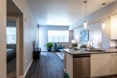 This thoughtfully designed single-floor residence offers a seamless blend of modern aesthetics and functional living. The open-concept layout connects the kitchen, dining, and living areas into one cohesive, light-filled space. Rich wood-look flooring flows throughout the main living areas, anchoring the contemporary design palette of soft gray walls and crisp white trim.

The kitchen is a standout feature, equipped with a full suite of stainless steel appliances including a refrigerator, range, and microwave. Sleek white cabinetry with clean-line hardware extends from floor to ceiling, complemented by gray countertops and under-cabinet lighting. A peninsula with bar seating serves as both a prep area and a casual dining space, while modern pendant lights add a refined touch overhead.

The living area is bright and welcoming, benefiting from large windows that invite ample natural light and offer views of surrounding greenery. A glass door provides direct access to the outdoors. The bedroom is a calm retreat, featuring plush carpeting, a generous closet, and windows with adjustable blinds for light control. The bathroom is clean and modern, outfitted with a single-sink vanity, a full mirror, and a shower with a curtain enclosure, all finished in a consistent, neutral palette.