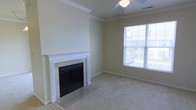 This single-floor residence offers a comfortable and well-appointed living space designed with classic details and a welcoming ambiance. The main living area is a standout feature, showcasing a traditional fireplace framed by an elegant white surround with decorative pilasters and a tile hearth, making it a natural focal point of the room. Soft, plush wall-to-wall carpeting runs throughout the space, adding warmth and comfort underfoot. Crown molding trims the upper edges of the walls, lending a refined architectural touch to the interior. Neutral, light-toned walls provide a clean and versatile backdrop throughout. A ceiling fan with a light fixture is centrally positioned, offering both air circulation and ambient lighting. Large windows allow generous amounts of natural light to pour into the living area, creating a bright and airy atmosphere while offering views of the surrounding exterior environment. The overall aesthetic is classic and understated, combining practical features with thoughtful design details to create an inviting, move-in-ready living environment suited to a variety of lifestyles.