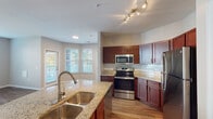 Located at 5741 Goodstone Dr in Raleigh, NC, this single-floor residence offers a well-designed layout with a cohesive blend of modern finishes and functional living spaces. The kitchen serves as a central highlight, showcasing rich cherry-toned cabinetry, granite countertops, a subway tile backsplash, and a full suite of stainless steel appliances including a range, microwave, and refrigerator. A peninsula with a double-basin sink and additional counter space enhances the kitchen's utility, while a bay window area brings in generous natural light. Hardwood-style flooring runs throughout the main living and kitchen areas, lending warmth and continuity to the open layout. The living area is spacious and bright, with a ceiling fan and easy access to adjacent rooms. Bedrooms are carpeted for comfort and feature ample natural light through sizable windows with blinds. Walk-in closets with built-in shelving are present in multiple bedrooms, providing excellent storage. The bathrooms are finished with rich wood vanities, large mirrors, and hardwood-style flooring. The primary bathroom includes a soaking tub, while a secondary bathroom offers a shower-tub combination. Overall, the space presents a clean, move-in-ready environment with practical amenities throughout.