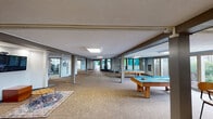 Located on The Royal Carriage Way in South Bend, Indiana, this expansive single-floor fitness and recreation facility offers a diverse range of amenities designed to support an active lifestyle. The centerpiece is a soaring indoor sports court with a dramatic vaulted ceiling supported by bold structural beams. Vibrant color-block walls in green, teal, orange, and navy energize the space, which features a full basketball court with hardwood-style flooring, running track lines, and abundant natural light streaming through large windows along the perimeter. Adjacent to the court, a dedicated cardio room houses treadmills, ellipticals, stationary bikes, and other machines, accented by motivational wall graphics. A separate strength training area provides free weights, a power rack, benches, and a dumbbell rack on durable rubber flooring. A yoga and stretching studio features soaring ceilings, a decorative tree-pose wall graphic, and rubber mat flooring for low-impact workouts. The facility also includes a spacious recreation lounge with multiple billiards tables and patterned carpet flooring, creating a relaxed social environment. Throughout, signage and wall art reinforce an energetic, motivational atmosphere, making this a well-rounded destination for both fitness and leisure.