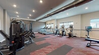 This fitness center offers a thoughtfully designed workout environment suited for a wide range of exercise routines. The space is divided into distinct functional zones, providing ample room for both cardio and strength training activities. 

The cardio zone is well-stocked with a row of Matrix treadmills, multiple elliptical trainers, and upright stationary bikes, all positioned to take advantage of natural light streaming in through several windows fitted with roller shades. A wall-mounted flat-screen TV keeps users entertained during workouts.

The strength training area features a multi-station cable machine, a rowing machine, a flat weight bench, and a fully stocked dumbbell rack with weights of varying sizes, catering to users of all fitness levels. Floor-to-ceiling mirrors line one wall, enhancing the sense of space and allowing users to monitor their form.

Underfoot, a durable patterned carpet in deep red and grey tones adds a contemporary touch while providing comfort and traction. Exposed industrial-style ductwork runs prominently along the ceiling, contributing a modern, urban aesthetic that contrasts with the clean, neutral-toned walls. Recessed lighting throughout ensures the space is well-lit at all times. The overall ambiance is functional, energetic, and welcoming.