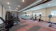 This fitness center offers a thoughtfully designed workout environment suited for a wide range of exercise routines. The space is divided into distinct functional zones, providing ample room for both cardio and strength training activities. 

The cardio zone is well-stocked with a row of Matrix treadmills, multiple elliptical trainers, and upright stationary bikes, all positioned to take advantage of natural light streaming in through several windows fitted with roller shades. A wall-mounted flat-screen TV keeps users entertained during workouts.

The strength training area features a multi-station cable machine, a rowing machine, a flat weight bench, and a fully stocked dumbbell rack with weights of varying sizes, catering to users of all fitness levels. Floor-to-ceiling mirrors line one wall, enhancing the sense of space and allowing users to monitor their form.

Underfoot, a durable patterned carpet in deep red and grey tones adds a contemporary touch while providing comfort and traction. Exposed industrial-style ductwork runs prominently along the ceiling, contributing a modern, urban aesthetic that contrasts with the clean, neutral-toned walls. Recessed lighting throughout ensures the space is well-lit at all times. The overall ambiance is functional, energetic, and welcoming.