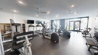 This fitness center offers a generously sized, single-floor exercise space designed to accommodate a comprehensive range of workout needs. The room features a durable, dark rubber flooring that spans the entire area, providing a safe and functional surface for all types of exercise.

The cardio section is well-appointed with multiple treadmills, elliptical trainers, a stationary bike, and a rowing machine, allowing users to engage in a full range of cardiovascular training. Strength training is supported by a dedicated free weights area, complete with a multi-tiered dumbbell rack, kettlebells, and weight plates, as well as select cable and resistance machines for targeted muscle work. A flat bench is also available for additional strength exercises.

Several wall-mounted flat-screen televisions are positioned throughout the room, ensuring visibility from various workout stations. Multiple ceiling fans are installed overhead, promoting airflow and comfort during intense sessions, while recessed lighting keeps the space well-lit throughout.

Large floor-to-ceiling windows and a set of glass double doors on one wall invite abundant natural light and offer views of the surrounding outdoor area. The overall design is modern and clean, with a neutral color palette of white and charcoal-grey accent walls that contributes to a focused and energizing atmosphere. Restroom or changing facilities appear to be accessible directly from the fitness floor via interior glass-panel doors.