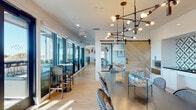 Located at 28168 Mission Blvd in Hayward, California, this single-floor amenity space delivers a refined blend of modern and transitional design. Light hardwood flooring runs throughout, creating a warm, cohesive foundation that connects each distinct zone seamlessly.

The main gathering area features soaring ceilings clad in white shiplap, anchored by a large, plush sectional sofa and a dramatic feature wall showcasing multiple flat-screen displays set against a bold, grid-patterned backdrop. Built-in shelving with dark navy cabinetry adds a sophisticated accent.

Adjacent to this lounge, a striking barn door with a herringbone wood panel design leads to a central corridor and kitchenette. The kitchenette is outfitted with sleek white cabinetry, dark stone countertops, a brass sink, and a bold geometric black-and-white tile backsplash that serves as a standout design element.

The dining area seats a generous number of guests at a long, dark wood communal table surrounded by woven bistro-style chairs. An industrial-modern chandelier with exposed Edison bulbs illuminates the space above. Floor-to-ceiling windows and glass doors flood the interior with natural light and provide direct access to an outdoor terrace, offering open views of the surrounding Hayward skyline and beyond.