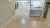 This single-floor space is designed with an open-concept layout that seamlessly connects the living area and kitchen, creating a bright and functional environment. The living area features light-toned hardwood flooring that extends throughout, contributing to a clean and airy atmosphere. Soft, neutral walls in a warm cream tone provide a versatile backdrop that complements the space's overall design.

A pendant light fixture is centered above the living area, providing ambient lighting, while the kitchen benefits from additional pendant lighting that enhances its functionality. The kitchen is outfitted with white cabinetry, white appliances including a refrigerator and stove, and a darker countertop that offers a subtle contrast. A breakfast bar or counter peninsula separates the kitchen from the living area, adding both a practical dining option and a natural visual division between the two spaces.

To the left, mirrored panels are mounted along the wall, reflecting light and adding a sense of depth to the room. The space is finished with white baseboards that frame the flooring cleanly. Overall, the layout is straightforward and well-organized, making efficient use of the available floor space.