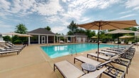 Located at 140 N Davis Rd in LaGrange, Georgia, this outdoor amenity space centers around a generously sized swimming pool with crystal-clear turquoise water. The pool is surrounded by an expansive concrete sun deck lined with cushioned lounge chairs and shaded umbrella stations, providing ample space for relaxation and sunbathing. A white pergola structure adjacent to the poolside offers a shaded retreat with stylish day beds, creating an intimate lounging area. Flanking the pool area are two distinct support buildings — one featuring large glass doors that open directly to the pool deck, ideal for an indoor-outdoor social space, and a second tan-sided utility or restroom facility. A fire pit area and additional seating clusters are visible throughout the deck, enhancing the social atmosphere. The perimeter is enclosed by a black iron fence, providing security while maintaining open views of the surrounding mature trees and lush greenery. The overall ambiance is that of an upscale, resort-inspired retreat, thoughtfully designed for outdoor recreation and community gathering in a well-maintained setting.