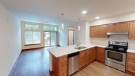 Located in Poulsbo, Washington, this single-floor residence at Viking Way NW offers a well-designed open-concept layout that blends functionality with comfortable living. The kitchen serves as a central highlight, featuring warm-toned wood cabinetry, crisp white countertops, and a center island equipped with a stainless steel sink and dishwasher. Additional stainless steel appliances include a freestanding range and a full-size refrigerator, while ample upper and lower cabinetry provides generous storage throughout. Pendant lighting above the island and recessed ceiling lights add layered illumination to the space.

The kitchen flows naturally into the living area, which is finished with hardwood-style flooring and benefits from abundant natural light through large windows and a sliding glass door that opens to a balcony overlooking lush greenery. A track lighting fixture further brightens the living space.

The bedrooms are carpeted for comfort and feature neutral tones, wood baseboards, and spacious closets. One bedroom includes a walk-in closet, while others offer sliding or hinged closet doors. Windows frame views of surrounding trees, enhancing the tranquil Pacific Northwest atmosphere throughout the unit.