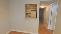 This single-floor residential space offers a clean, functional layout with a warm and neutral aesthetic throughout. The living area features smooth hardwood-style flooring that extends across the main areas, lending a cohesive and inviting feel to the space. A standout architectural detail is the built-in pass-through window that connects the living area to the kitchen, framed neatly within the wall and accented with a white ledge that could serve as a convenient serving counter or casual dining surface. Through the pass-through, the kitchen reveals white cabinetry offering ample storage, a full-size refrigerator, and overhead flush-mount lighting that keeps the space bright and well-illuminated. The walls are finished in a soft, neutral tone that complements the light flooring and white trim work, creating an airy and clean atmosphere. A hallway extends beyond the main living area, leading to additional rooms and providing a sense of depth and connectivity within the floor plan. Flush-mount ceiling fixtures provide consistent lighting throughout. Clean baseboards and crisp trim details reflect careful attention to finish work, making this a well-maintained and move-in ready space suitable for comfortable everyday living.