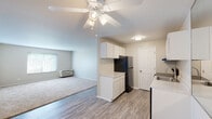 This single-floor unit offers a practical and well-designed layout that connects the kitchen and living areas in an open, flowing arrangement. The kitchen is equipped with black stainless appliances, including a refrigerator, electric range, and dishwasher. White cabinetry provides ample storage, while a peninsula with a built-in sink and countertop serves as both a functional workspace and a natural divider between the kitchen and living space. Light wood-style flooring runs throughout the kitchen and entry areas, transitioning to plush carpeting in the spacious living room. The living room is generously sized, featuring a ceiling fan, a wall-mounted air conditioning unit, and a window that allows natural light to filter in. A hallway leads to additional rooms, including a bathroom fitted with a white vanity, large mirror, and combination tub and shower. Neutral tones throughout the unit — from the soft beige walls to the light countertops — create a clean, cohesive aesthetic. The overall layout is efficient and open, making the space feel bright and easy to navigate.