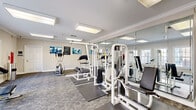 This fitness center is a thoughtfully designed, single-level space that accommodates a comprehensive range of workout equipment within a clean, organized layout. The room features patterned carpet tile flooring with rubber exercise mats positioned beneath the equipment, providing both durability and comfort during workouts. Crown molding along the ceiling adds a refined architectural detail, while flush-mounted fluorescent light panels ensure consistent, bright illumination throughout the space.

A defining visual feature is the large motivational typography mural covering one wall, displaying fitness-related terms such as "Exercise," "Cardio," "Strength," and "Wellness" in bold blue and white lettering. Full-length mirrors line an adjacent wall, visually expanding the room and allowing users to monitor their form during exercise.

The equipment selection includes multiple cable-and-weight strength machines, a treadmill, an elliptical trainer, and a seated abdominal machine. A wall-mounted flat-screen television and a hand sanitizer station are also present. Multiple windows along one wall invite natural light, contributing to an open, energizing atmosphere. Safety amenities include a prominently placed AED unit and posted fitness center hours, reflecting a well-maintained, user-focused environment.