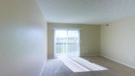 This single-floor space offers a clean, well-lit environment designed for comfortable everyday living. The main living area is bathed in natural light, courtesy of a large sliding glass door fitted with vertical blinds that allow for adjustable light control throughout the day. The door opens directly onto a private balcony, which overlooks a lush, green landscape — providing a peaceful outdoor retreat and a refreshing connection to nature.

The interior features neutral, warm-toned walls that complement the light carpeted flooring, creating a soft and welcoming ambiance throughout. The smooth ceiling adds to the open, airy feel of the room, while the understated color palette provides a versatile backdrop suited to a variety of furnishing styles.

The layout is straightforward and functional, with generous wall space that lends flexibility for furniture arrangement. The combination of abundant natural light, outdoor access, and a calm, neutral aesthetic makes this a comfortable and practical space. Whether used as a relaxing retreat or a day-to-day living environment, the space balances simplicity with a bright, inviting character.