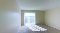 This single-floor space offers a clean, well-lit environment designed for comfortable everyday living. The main living area is bathed in natural light, courtesy of a large sliding glass door fitted with vertical blinds that allow for adjustable light control throughout the day. The door opens directly onto a private balcony, which overlooks a lush, green landscape — providing a peaceful outdoor retreat and a refreshing connection to nature.

The interior features neutral, warm-toned walls that complement the light carpeted flooring, creating a soft and welcoming ambiance throughout. The smooth ceiling adds to the open, airy feel of the room, while the understated color palette provides a versatile backdrop suited to a variety of furnishing styles.

The layout is straightforward and functional, with generous wall space that lends flexibility for furniture arrangement. The combination of abundant natural light, outdoor access, and a calm, neutral aesthetic makes this a comfortable and practical space. Whether used as a relaxing retreat or a day-to-day living environment, the space balances simplicity with a bright, inviting character.