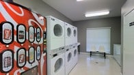 This single-floor communal laundry facility is designed with practicality and convenience in mind. The room features multiple stacked washer and dryer units lined along one wall, providing ample laundry capacity for residents or guests. The machines are arranged in a neat, accessible configuration, maximizing the use of the available floor space.

The interior is finished with neutral gray walls and light-toned flooring, creating a clean and utilitarian atmosphere. Bright fluorescent overhead lighting ensures the space is well-lit throughout. A window fitted with horizontal blinds allows for natural light while maintaining privacy.

A colorful beverage vending machine is positioned near the entrance, offering refreshments while patrons wait. Seating is available along the far wall, with a pair of white molded chairs and a bench, providing a comfortable spot to rest during laundry cycles.

Overall, the space is thoughtfully organized to deliver a straightforward and efficient laundry experience, with all essential amenities within easy reach.