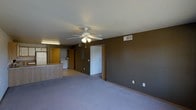 This single-floor space presents an open-concept layout that combines the living area with a partially visible kitchen and utility zone. The living area is finished with wall-to-wall gray carpeting, providing a soft and comfortable underfoot feel throughout the space. Earthy brown-painted walls lend a warm, grounded tone to the interior, complemented by white ceilings that help brighten the room.

A white ceiling fan with an integrated light fixture is centrally mounted, offering both air circulation and ambient lighting to the main living space. HVAC vents are positioned along the upper walls, ensuring efficient climate control throughout the area.

Toward the back of the space, an open passageway leads to an adjoining area housing a washer and dryer, indicating dedicated in-unit laundry functionality. Wooden cabinetry is visible in the kitchen zone, which is partially separated from the living area by a low counter or partition, maintaining a sense of openness while defining the functional cooking space. A doorway visible from the living area provides access to additional rooms. The overall layout is practical and straightforward, making efficient use of the available single-floor footprint.
