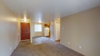 This single-floor residential unit offers a straightforward and functional layout designed for comfortable everyday living. The main living area greets you upon entry through a solid wood front door, and is finished with plush carpeting that adds warmth underfoot. Neutral beige walls throughout create a clean, cohesive look that complements a variety of furnishing styles. The space transitions smoothly from the carpeted living area to hard-surface flooring as you move toward the kitchen, which is outfitted with wood-toned cabinetry and includes a full-size refrigerator. A window in the kitchen area allows for natural light to filter through. A baseboard heating unit along the far wall ensures consistent warmth during cooler months. Adjacent to the main living space, a set of closet doors provides convenient storage solutions. Ceiling-mounted light fixtures provide ambient lighting throughout. The textured ceiling adds a subtle design detail consistent across the unit. Overall, the space presents a clean, move-in-ready environment with a practical and unpretentious character suited to a wide range of residents.
