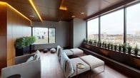 This thoughtfully designed single-floor space combines contemporary minimalism with warm, organic materials to create a refined and calming environment. Rich hardwood flooring runs throughout, complemented by full-height wood-paneled walls that lend a sense of warmth and continuity. The ceiling is a standout architectural feature, composed of layered wood panels with integrated amber cove lighting that casts a soft, golden glow across the room, enhancing the overall ambiance.

The lounge area is furnished with plush, neutral-toned sofas and chaise seating arranged to encourage relaxation and social interaction. Sculptural stone accent pieces and slender brass side tables punctuate the space with subtle elegance. A marble-clad feature wall houses a linear fireplace, adding a focal point of luxury to one end of the room.

Large windows on multiple sides flood the interior with natural light and frame impressive urban skyline views, with built-in planters along the window ledges introducing a refreshing touch of greenery. A beverage station with warm wood cabinetry and white countertops provides practical amenities. Additional potted plants near the windows reinforce the biophilic design approach, creating a serene atmosphere that balances sophistication with comfort.