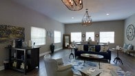 This well-appointed single-floor space showcases a thoughtfully designed open layout that seamlessly blends living and dining areas into one cohesive environment. The interior is anchored by two striking decorative chandeliers — one featuring an ornate cage-style design and another with a rope-accented frame — that add a touch of refined character to the space. Smooth, neutral gray walls complement the rich, dark hardwood-style flooring, creating a warm yet contemporary aesthetic throughout. Multiple windows dressed with clean white horizontal blinds invite generous natural light, brightening the interior and enhancing its sense of openness. The living area is furnished with a deep navy blue sofa, a coordinating accent chair in soft gray with nailhead trim, and a patterned area rug in blue and white tones that adds texture and visual interest. A round side table and an oval coffee table complete the seating arrangement. Toward the back, a dining area is visible with additional seating, a full-length mirror with a wood frame, and tasteful wall décor. The overall ambiance is modern and inviting, balancing casual comfort with stylish design elements that make this space both functional and visually appealing.