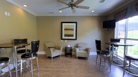 This single-floor space offers a well-appointed lounge area designed for comfort and casual interaction. The room features warm beige walls complemented by crisp white crown molding and baseboards, creating a clean, polished aesthetic throughout. The flooring consists of cork or stone-look tile, adding a natural, grounded feel underfoot.

Seating is thoughtfully arranged to encourage conversation, with a pair of cushioned armchairs positioned around a low cylindrical accent table at the room's center. Flanking the space are tall bar-height chairs with black leather upholstery and chrome frames, paired with a high counter along the perimeter — offering an alternative seating option for those preferring an elevated vantage point.

A ceiling fan with integrated lighting is centrally mounted, providing both air circulation and ambient illumination alongside recessed ceiling lights. A framed landscape artwork serves as a focal point on the main wall, while a wall-mounted flat-screen television is visible to the side, adding a functional media element. The overall ambiance is relaxed yet refined, making the space well-suited for socializing or informal gatherings.