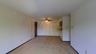 This single-floor space features an open-concept living area that flows seamlessly toward a partially visible kitchen. The main living area is finished with light-toned flooring that lends a clean, neutral aesthetic throughout the space. White walls keep the interior bright and versatile, providing a blank canvas for personalization.

A ceiling fan with an integrated light fixture is centrally mounted, offering both ventilation and ambient lighting to the main living area. Dark baseboards provide a subtle contrast against the light walls and flooring, adding a modest degree of visual definition to the room.

Toward the rear of the living space, a kitchen counter is partially visible, suggesting a connected or open kitchen layout. Dark cabinetry overhead adds a contrasting accent to the otherwise light interior. A door with a decorative ventilation panel is positioned along the back wall, providing access to an adjacent area or exterior.

Electrical outlets and light switches are distributed along the walls, ensuring functional utility throughout. Overall, the space presents a straightforward, functional layout with a neutral palette well-suited for a variety of furnishing styles.