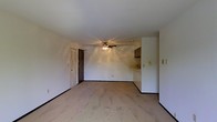 This single-floor space features an open-concept living area that flows seamlessly toward a partially visible kitchen. The main living area is finished with light-toned flooring that lends a clean, neutral aesthetic throughout the space. White walls keep the interior bright and versatile, providing a blank canvas for personalization.

A ceiling fan with an integrated light fixture is centrally mounted, offering both ventilation and ambient lighting to the main living area. Dark baseboards provide a subtle contrast against the light walls and flooring, adding a modest degree of visual definition to the room.

Toward the rear of the living space, a kitchen counter is partially visible, suggesting a connected or open kitchen layout. Dark cabinetry overhead adds a contrasting accent to the otherwise light interior. A door with a decorative ventilation panel is positioned along the back wall, providing access to an adjacent area or exterior.

Electrical outlets and light switches are distributed along the walls, ensuring functional utility throughout. Overall, the space presents a straightforward, functional layout with a neutral palette well-suited for a variety of furnishing styles.