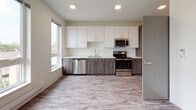 This modern single-floor unit offers a clean, contemporary design with a cohesive aesthetic carried throughout each room. Wood-look plank flooring runs continuously across the space, providing warmth and visual continuity.

The kitchen is a standout feature, outfitted with a full suite of stainless steel appliances including a refrigerator, electric range, over-the-range microwave, and dishwasher. Two-tone cabinetry pairs crisp white upper cabinets with wood-grain lower cabinets, complemented by light quartz countertops and a stainless steel undermount sink with a modern faucet. Recessed lighting and a large window bathe the kitchen in natural light, enhancing its open and functional feel.

The bedroom is a well-proportioned, neutral-toned room featuring a wall-mounted mini-split air conditioning unit for individual climate control. The space is bright and clean, with modern door hardware providing a subtle contemporary touch.

The bathroom is thoughtfully designed with a white vanity offering ample drawer storage, a large mirror with above-mount lighting, and a toilet. Grab bars are present, indicating accessibility-conscious design, and the light wood-look tile flooring ties the space back to the rest of the unit.

Overall, the unit presents a fresh, modern interior with quality finishes and practical amenities throughout.