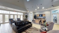 This single-floor space offers an open-concept layout that seamlessly integrates a lounge area, kitchenette, and dining space into one cohesive, multifunctional environment. The vaulted ceilings with a skylight create an impressive sense of vertical space, while recessed lighting and a ceiling-mounted pendant fixture ensure the interior remains well-lit throughout.

The lounge area is furnished with a large black leather sectional sofa and complementary seating, arranged atop a patterned area rug over light-toned tile flooring. A wall-mounted television serves the seating area, and large French-style glass doors along one wall flood the space with natural light while offering views of a lush outdoor area.

The kitchenette, positioned toward the rear, features light wood cabinetry, granite-style countertops, a black refrigerator, and a mounted television above the counter. Industrial-style metal bar stools line the counter, providing casual seating. An adjacent dining area with table and chairs rounds out the space. The overall ambiance is modern and welcoming, making it well-suited for social gatherings and communal use.