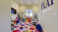 This cheerful children's playroom is designed to inspire creativity and active play in a welcoming, well-appointed space. The room is anchored by a bold, multicolored geometric-patterned carpet that immediately sets a lively and energetic tone. Soft sage-green walls contribute a calm backdrop that complements the vivid furnishings and décor throughout.

The space is thoughtfully organized into multiple activity zones. A central open area provides ample room for imaginative play, featuring a dollhouse, soft foam climbing blocks, and child-sized tables and chairs. Wall-mounted shelving units display a curated selection of toys, books, and educational materials, keeping everything accessible and organized. A play kitchen and infant activity stations cater to a range of age groups.

A large, multi-pane window along one wall floods the room with natural light and offers views of the surrounding greenery, enhancing the open and airy feel of the space. Recessed ceiling lighting and a decorative cage-style chandelier provide warm, even illumination throughout. A wall-mounted flat-screen television is positioned in one corner for additional entertainment options.

The entry area transitions seamlessly from wood-look luxury vinyl plank flooring into the play space, with glass-paneled doors providing a bright, welcoming arrival. Inspirational wall art and bold "PLAY" signage reinforce the room's playful and inclusive character.